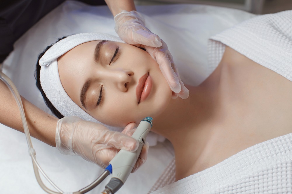 Aesthetic treatment session featuring a woman receiving a facial procedure with a handheld device, performed by a professional in a spa setting. The client has her eyes closed and appears relaxed, while wearing a headband and a robe.