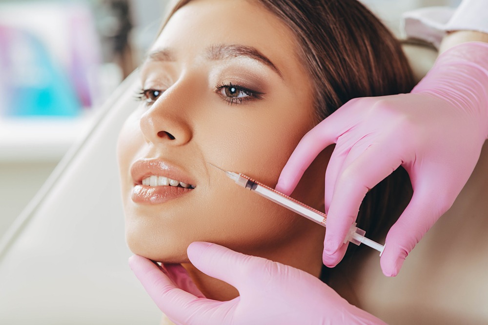 Close-up of a woman receiving a cosmetic injection in her cheek, administered by a professional wearing pink gloves. The setting appears to be a modern aesthetic clinic, focusing on beauty treatments.