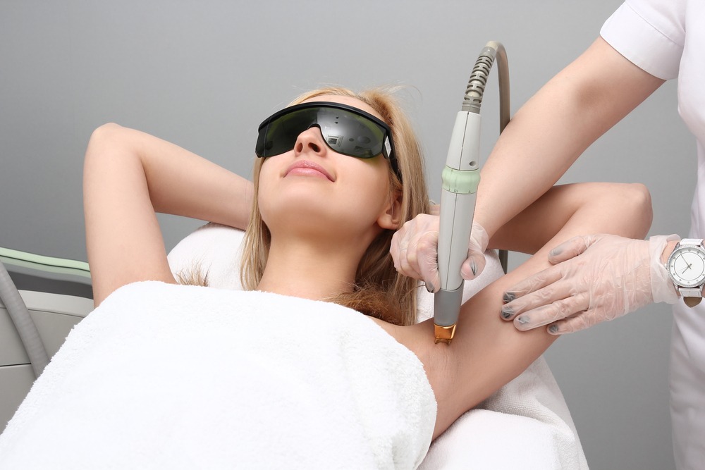 Woman receiving laser hair removal treatment in a professional clinic, wearing protective glasses and a towel. A technician operates the laser device on her underarm, showcasing modern aesthetics and skincare procedures.