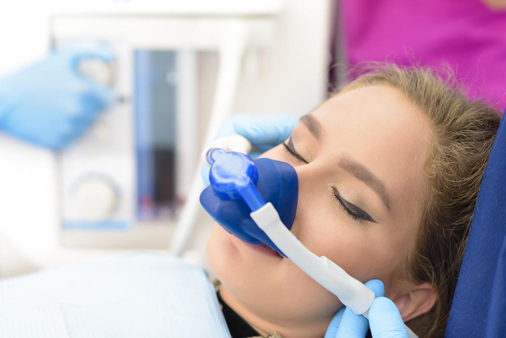 Patient receiving nitrous oxide sedation in a dental clinic, with a dental professional monitoring equipment in the background. The patient appears relaxed with eyes closed, wearing a nasal mask for sedation.