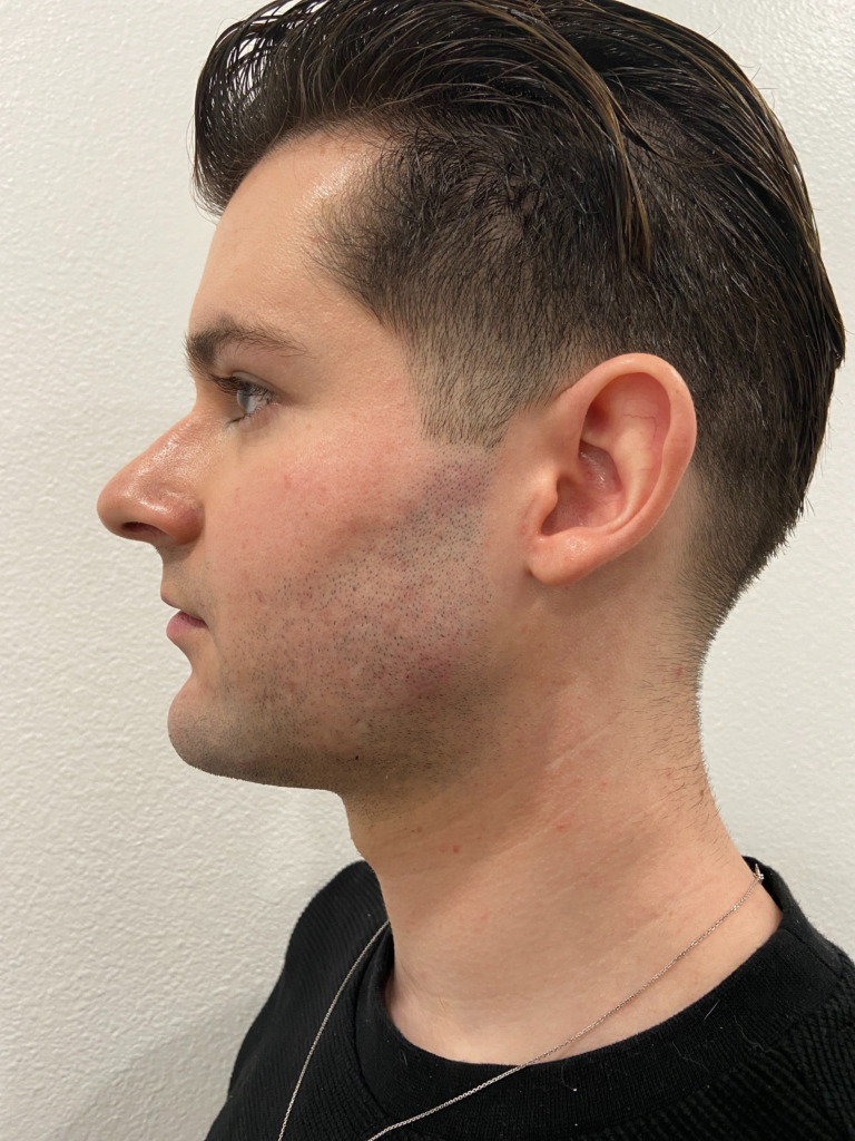 Profile view of a young man with short, styled hair and a subtle beard, wearing a black sweater and a silver necklace, against a plain white background.