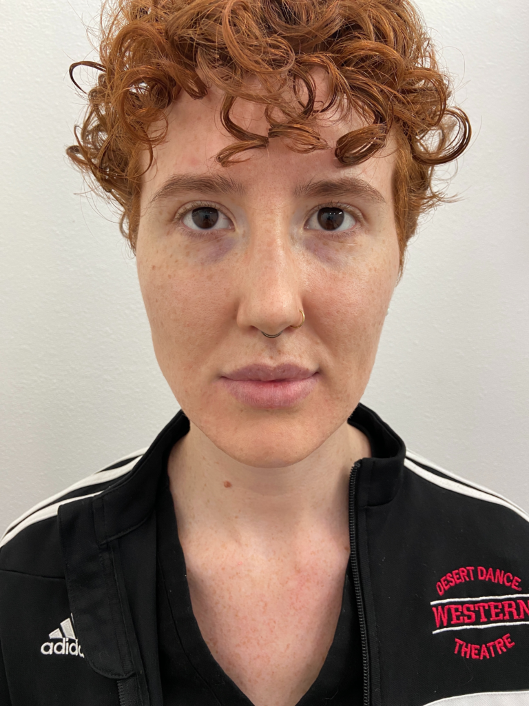 Portrait of a person with curly red hair and a nose ring, wearing a black jacket with 