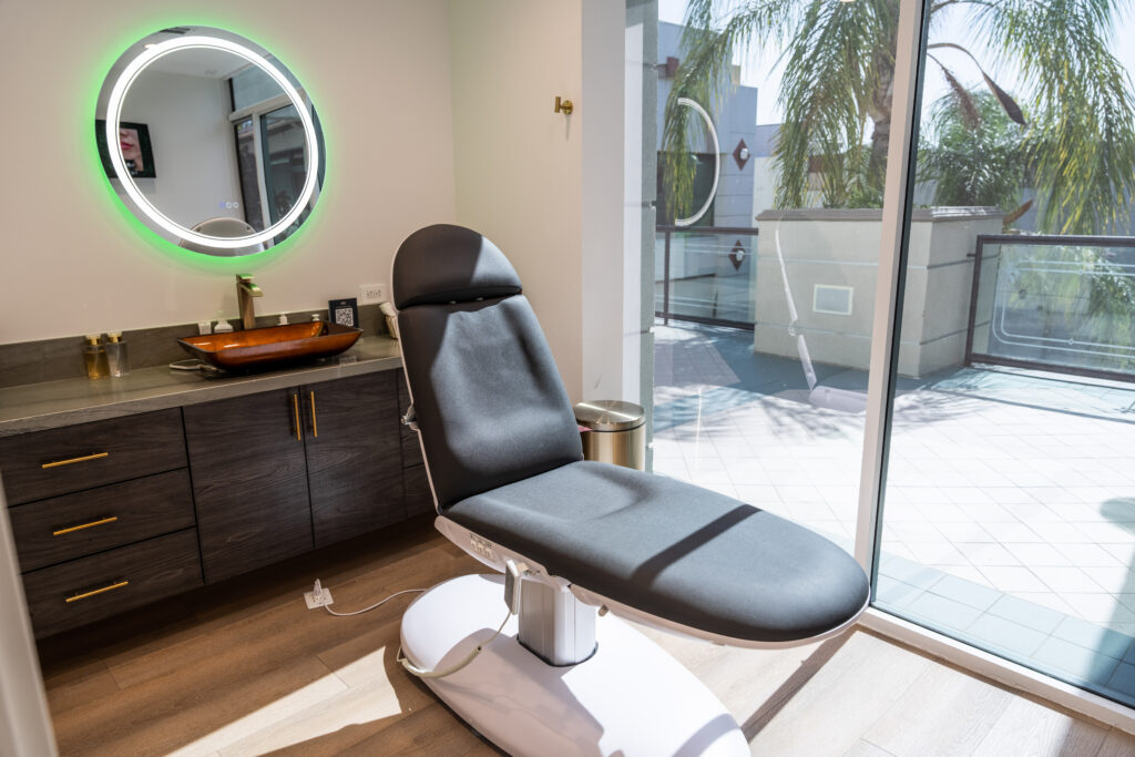 Modern aesthetic treatment room featuring a comfortable, adjustable treatment chair, stylish vanity with a wooden bowl, and a circular illuminated mirror. Large windows provide natural light and a view of the outdoor area with palm trees.