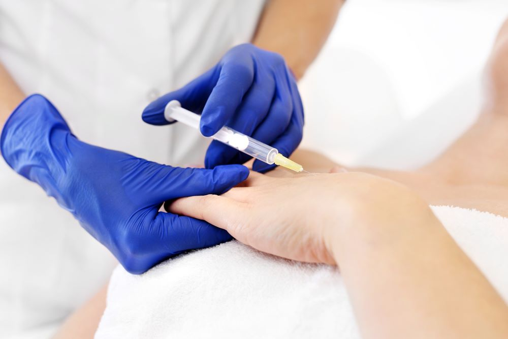 Healthcare professional administering an injection to a patient's hand, wearing blue gloves in a clinical setting.