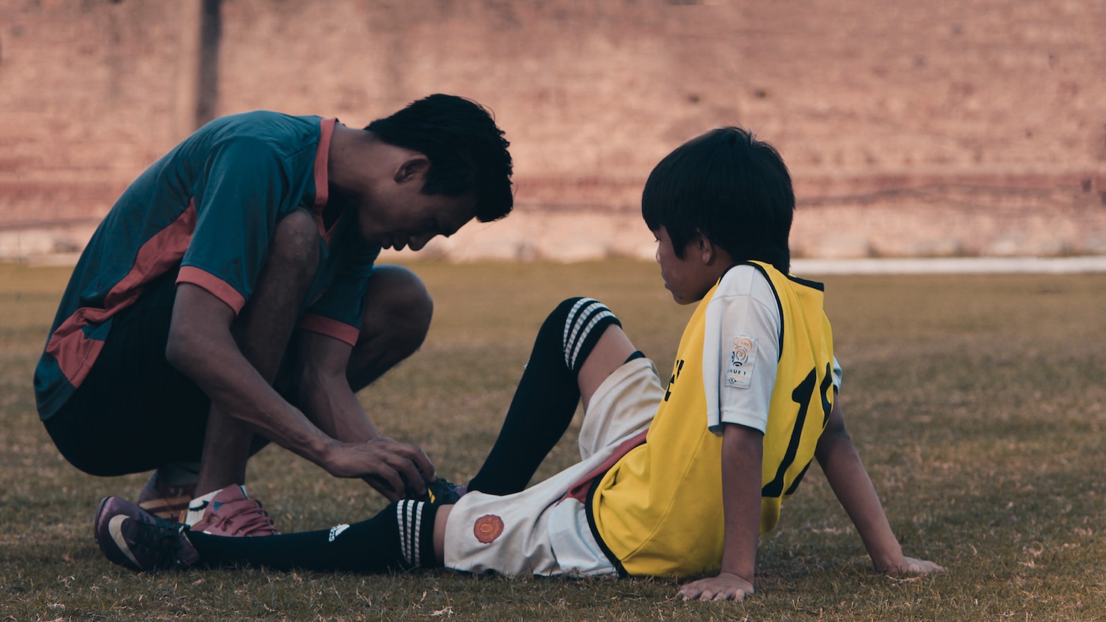A young boy in a yellow soccer jersey sits on the grass while an older male coach helps him adjust his soccer socks. The scene captures a moment of mentorship and preparation for a soccer game.